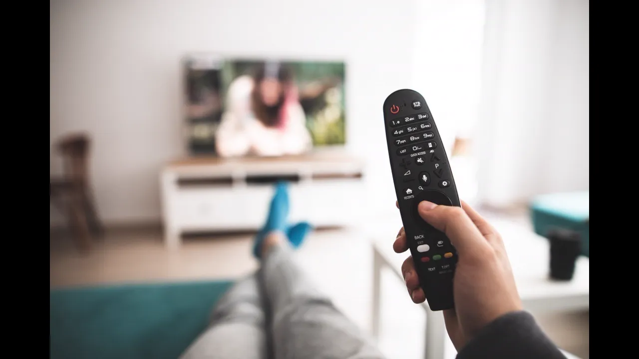A hand holding a TV remote pointed at a television in a bright living room