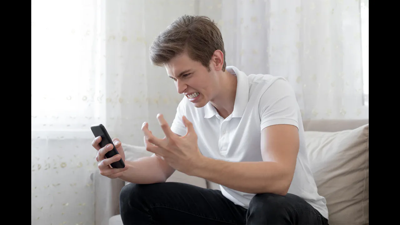 A frustrated man on a sofa holding a smartphone and gesturing at its blank screen