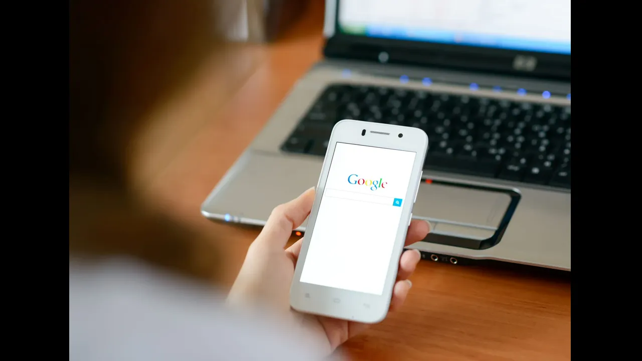 A hand holding a white smartphone showing a search page, with a laptop behind it